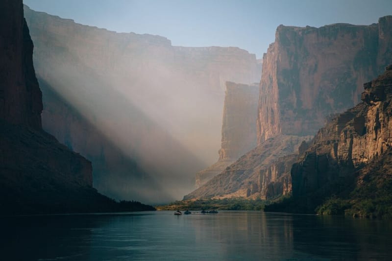 Sunbeams breaking through haze over the Colorado River with small rafts between towering Grand Canyon cliffs