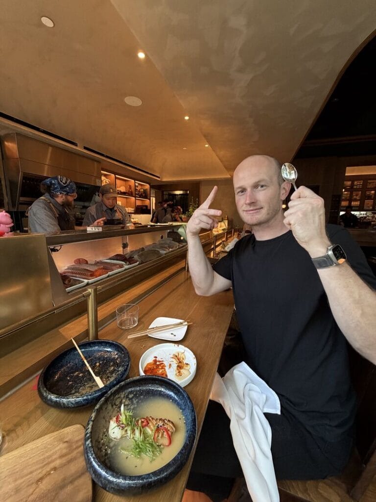 Bar Bruhis, a bald man in a black t-shirt, holding up a spoon at a sushi counter with bowls of soup and small dishes in front of him